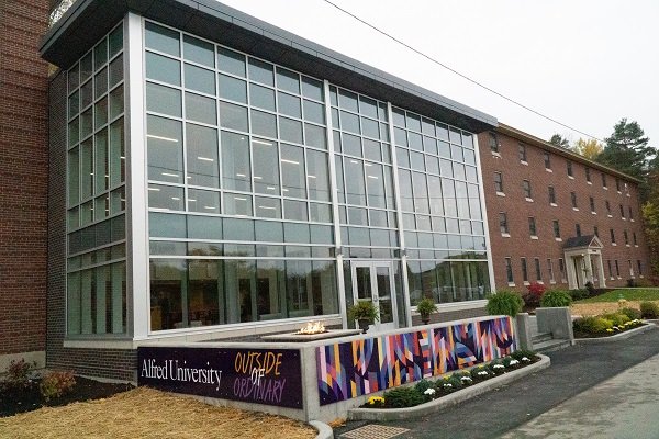 Glass building extension of Alfred University with a banner reading "Outside of Ordinary," adjacent to a brick structure. Overcast sky.