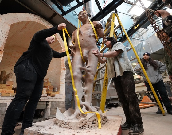 Students work together to support and measure a large clay sculpture of a standing figure inside a ceramics studio, using straps and a hoist system during the installation or firing process.