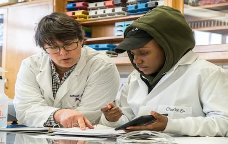 Two individuals wearing white lab coats collaborate at a table in a laboratory or classroom. One person points to a document or notebook, while the other holds a smartphone and a pen. Shelves in the background are stocked with scientific supplies and equipment, reinforcing the setting's academic and research focus.