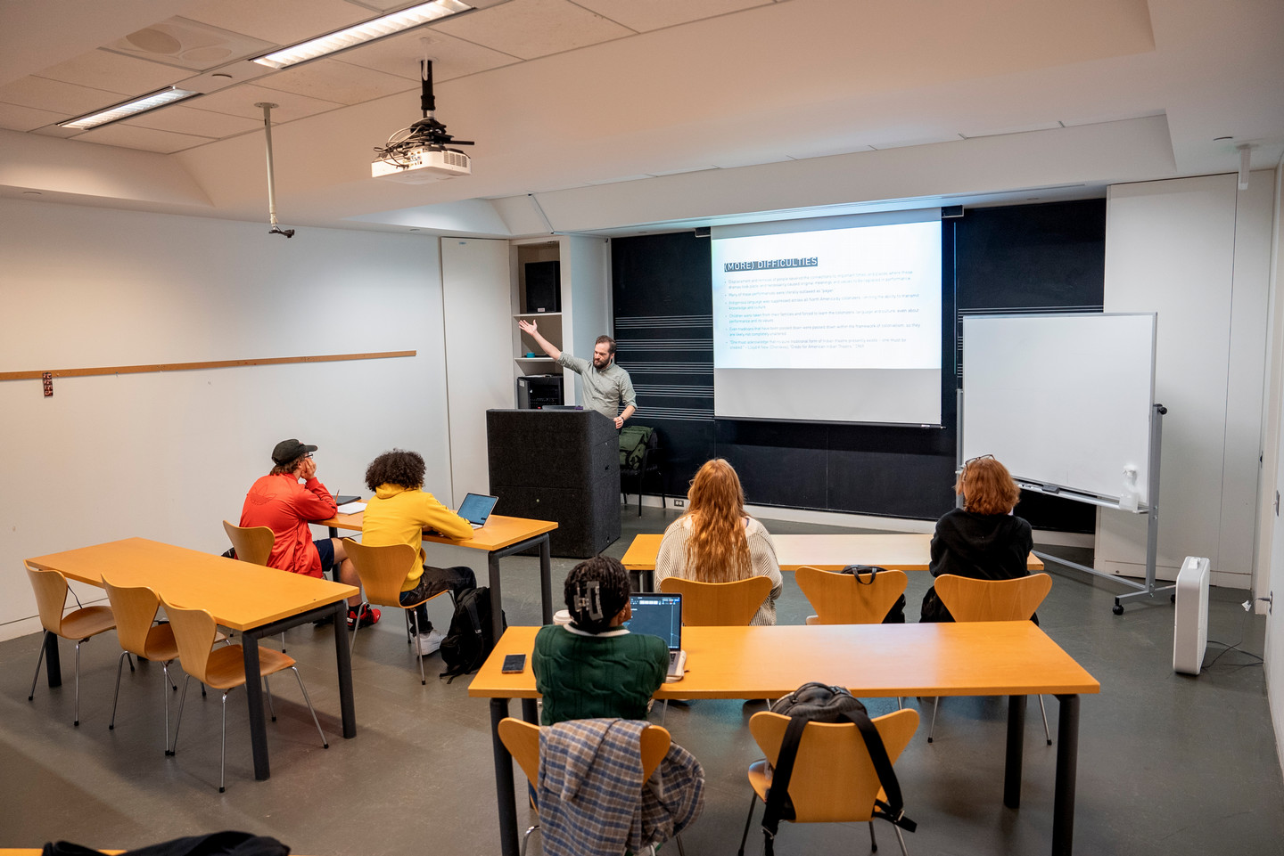 A lecturer stands at a podium, gesturing towards a projected slide. Five students, seated at desks with laptops, listen attentively. The setting is a modern classroom, conveying a focused and engaging learning environment.