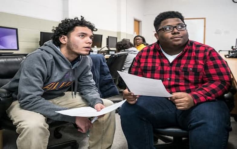 Two casually dressed individuals sit in a computer lab or classroom, discussing papers they’re holding. One wears a gray hoodie and beige pants, while the other wears a red and black plaid shirt with blue jeans. Behind them, others work at computer stations, and a whiteboard is visible on the wall, suggesting a collaborative academic environment.