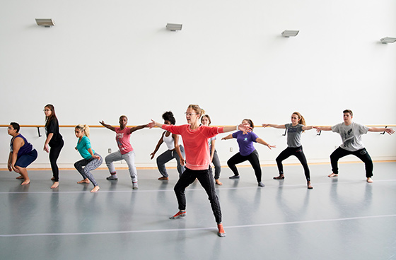 A dance instructor leads a group of students through movement exercises in a bright studio, with the class mirroring the instructor’s pose.