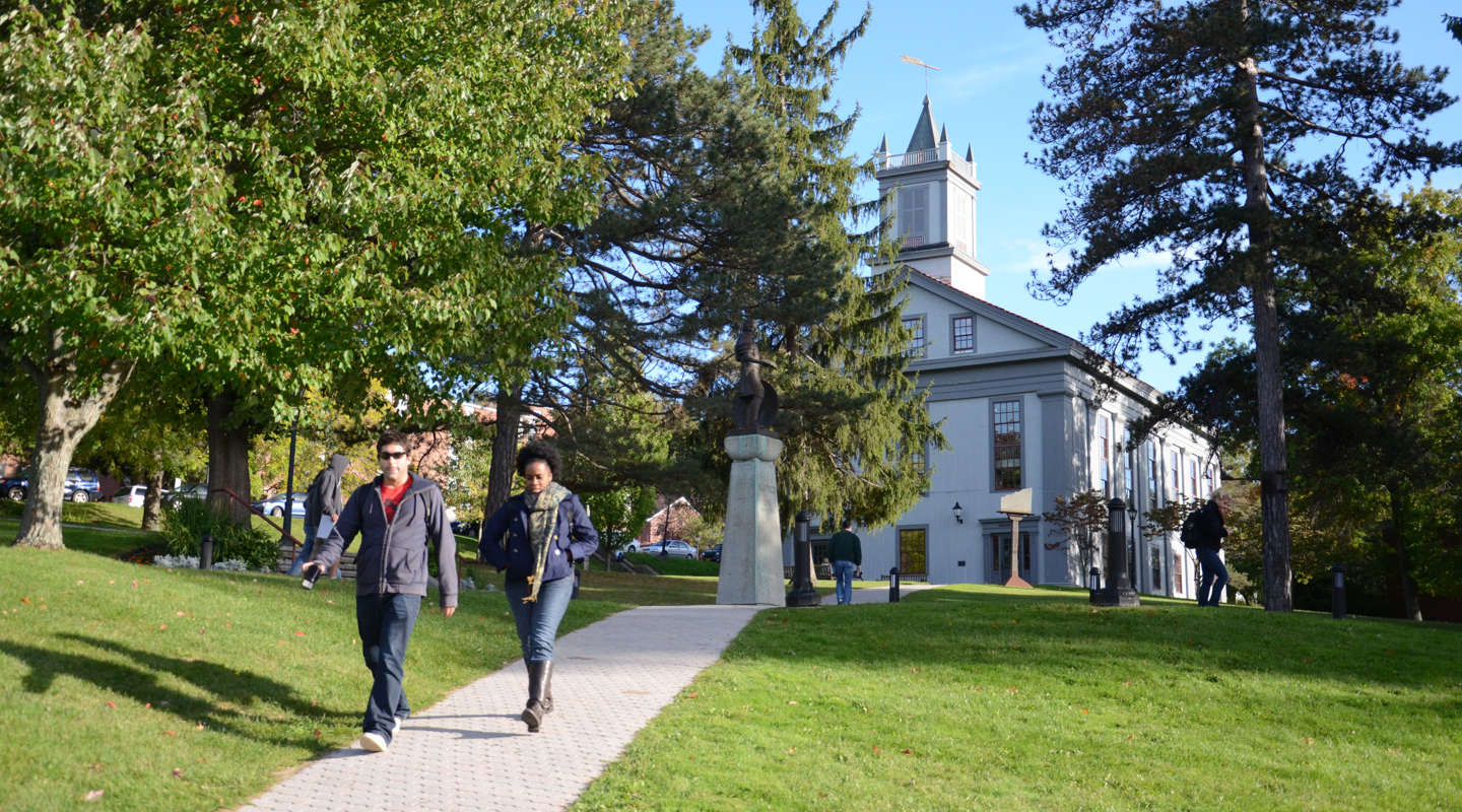 Students walk along a tree-lined path on the Alfred University campus with Alumni Hall visible in the background on a sunny day.