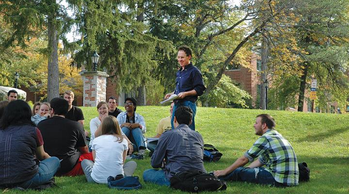A professor leads a class discussion with students seated in a circle on the lawn outside on a sunny day.