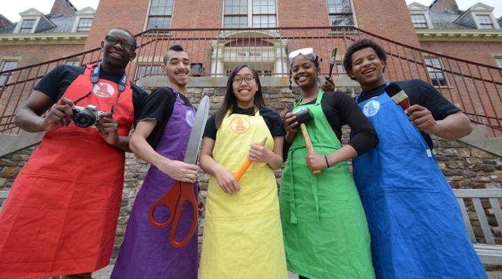 Five Alfred University students wearing colorful aprons and holding art tools while smiling in front of a campus building.