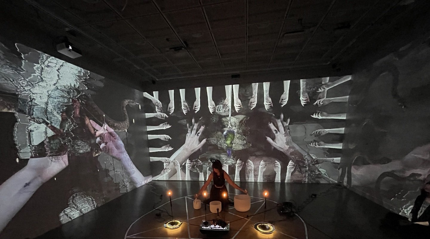 A dark room with immersive projection art on walls, depicting hands and abstract organic shapes. A person stands surrounded by circular lights, creating a mysterious, contemplative atmosphere.