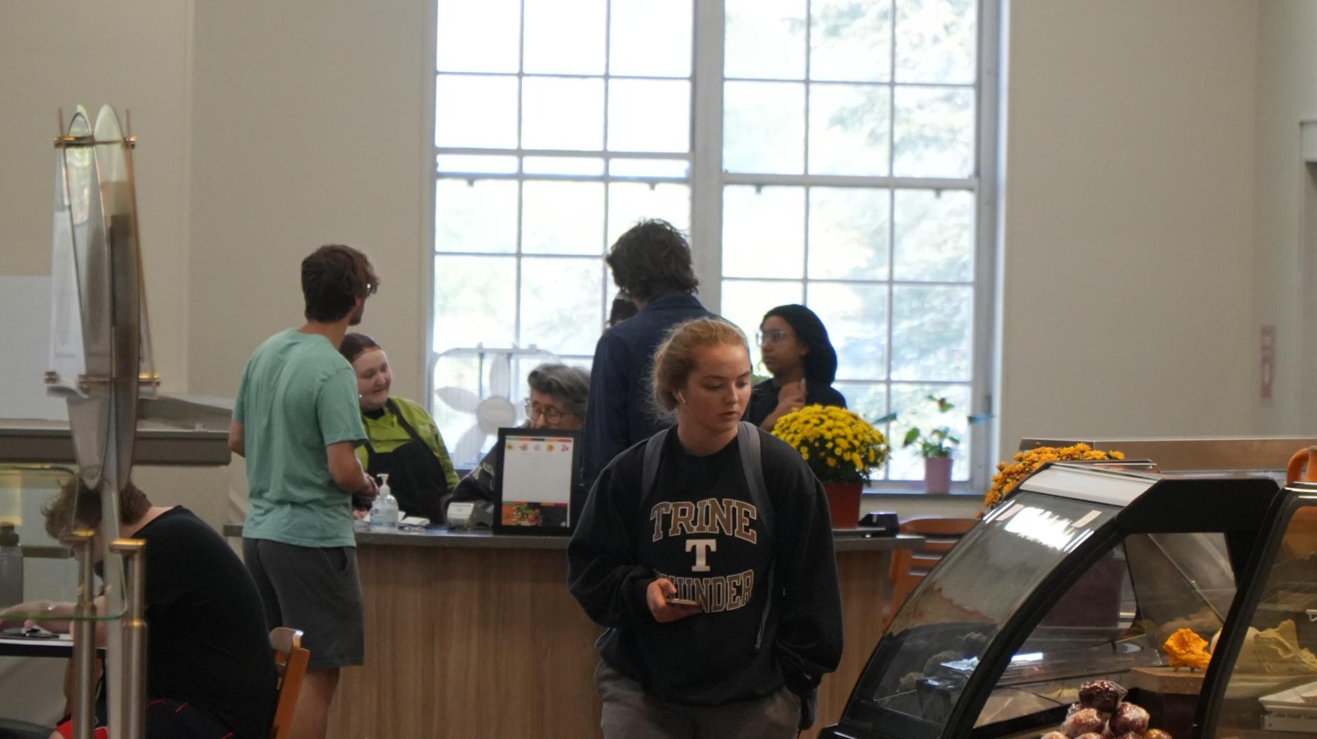 Several people stand and interact near a counter in a cafe or cafeteria; a woman in a Trine Thunder sweatshirt walks past a pastry display case in the foreground.
