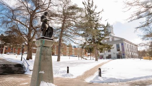 A statue of a King Alfred stands in the snow, with Alumni Hall visible in the background on Alfred University's campus.