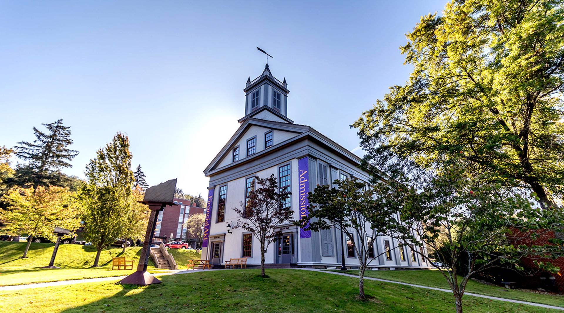 A historic building, Alumni Hall, with a tall steeple and banners reading "Admissions" and "Financial Aid" set amidst green lawns and trees under a clear blue sky. Bright and welcoming.