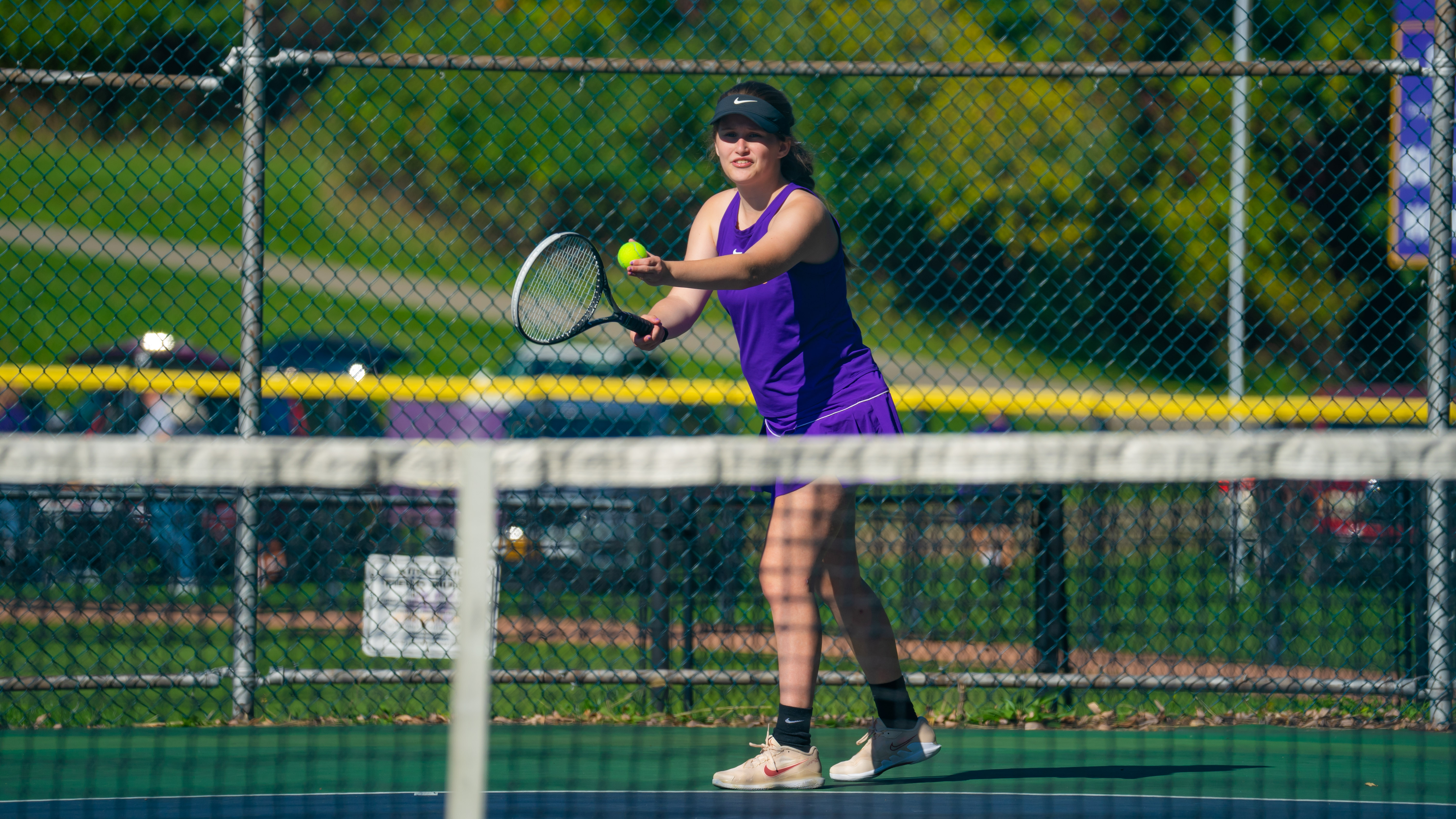 Female tennis player in purple uniform preparing to serve during a match on an outdoor court.