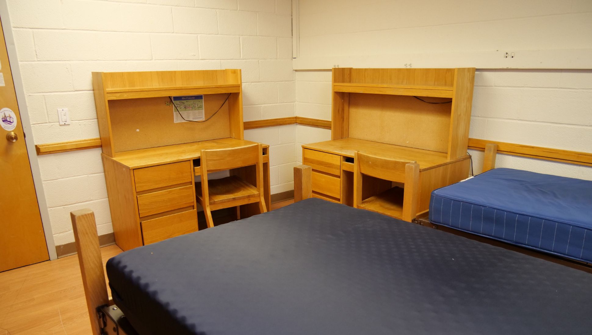 A college dorm room with two wooden desks and chairs, each featuring a built-in hutch and drawer storage. Two twin beds with blue mattresses are visible, one in the foreground and one to the side. The room has light-colored cinder block walls, a wooden floor, and simple wall-mounted outlets and lighting strips. A door with a small whiteboard and university sticker is partially visible on the left.