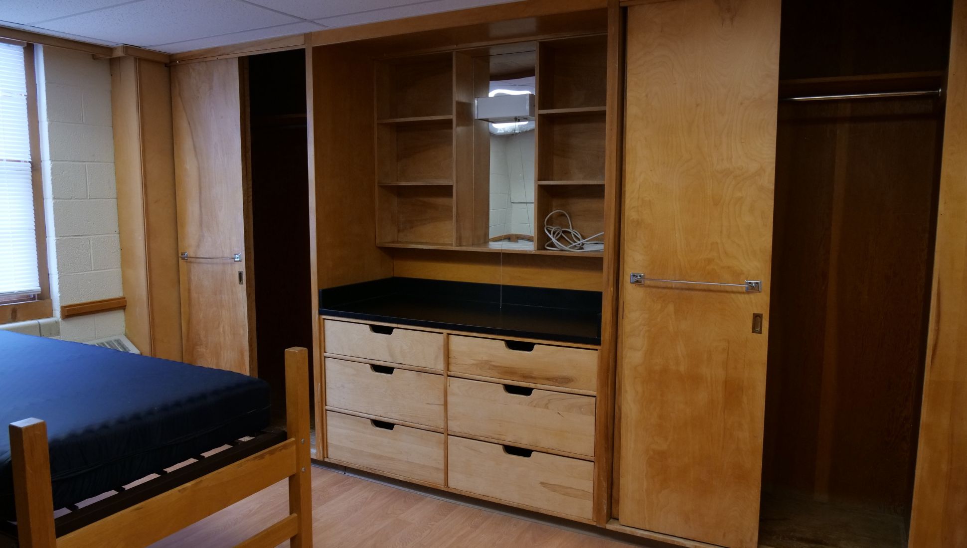A dorm room storage area with built-in wooden furniture, including two closets with sliding doors and a central vanity unit. The vanity has six drawers, open shelving, a mirror, and a black countertop. A bed frame with a blue mattress is partially visible in the foreground, and a window with blinds is on the left side of the image.