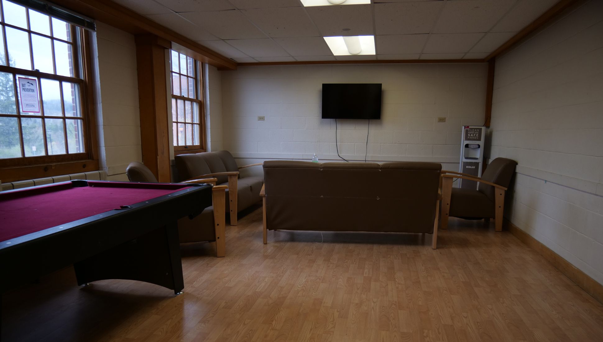A dorm common room with wooden floors, a wall-mounted TV, and brown cushioned seating including two armchairs and a couch arranged around the TV. A red pool table is in the foreground on the left side. Large windows line the left wall, allowing natural light into the room. A hand sanitizer dispenser labeled “Clean Safe Hands” is placed near the back wall. A sign on the window mentions “Bat Prevention.”