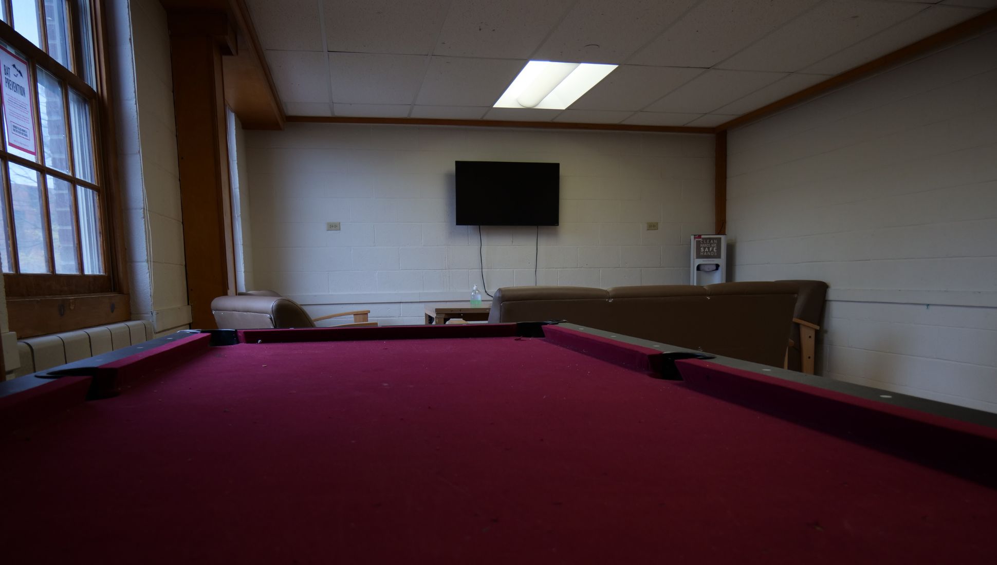 A common room with a red felt pool table in the foreground, facing a wall-mounted TV. Brown sofas and armchairs are arranged around the TV area, and a hand sanitizer dispenser labeled "Clean Safe Hands" stands against the wall to the right. The room has white cinder block walls, a drop ceiling with fluorescent lighting, and large wooden-framed windows on the left side. A sign on the window reads "Bat Prevention."