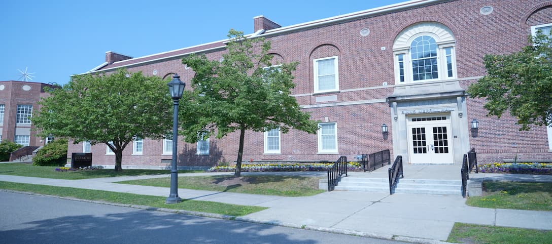 A large brick building with white-framed windows and a central entrance featuring double glass-paneled doors. Two trees flank the entrance, and a black lamppost stands near the sidewalk. Steps with black railings lead up to the entrance, surrounded by neatly trimmed grass and flower beds.