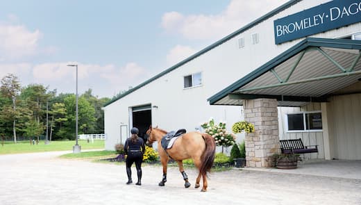 A person in equestrian gear leads a saddled brown horse toward the entrance of a large white building labeled "Bromeley-Daggett Equestrian Center." The building features an overhang supported by stone pillars and is surrounded by greenery and flowers. The sky is partly cloudy, with trees visible in the background.