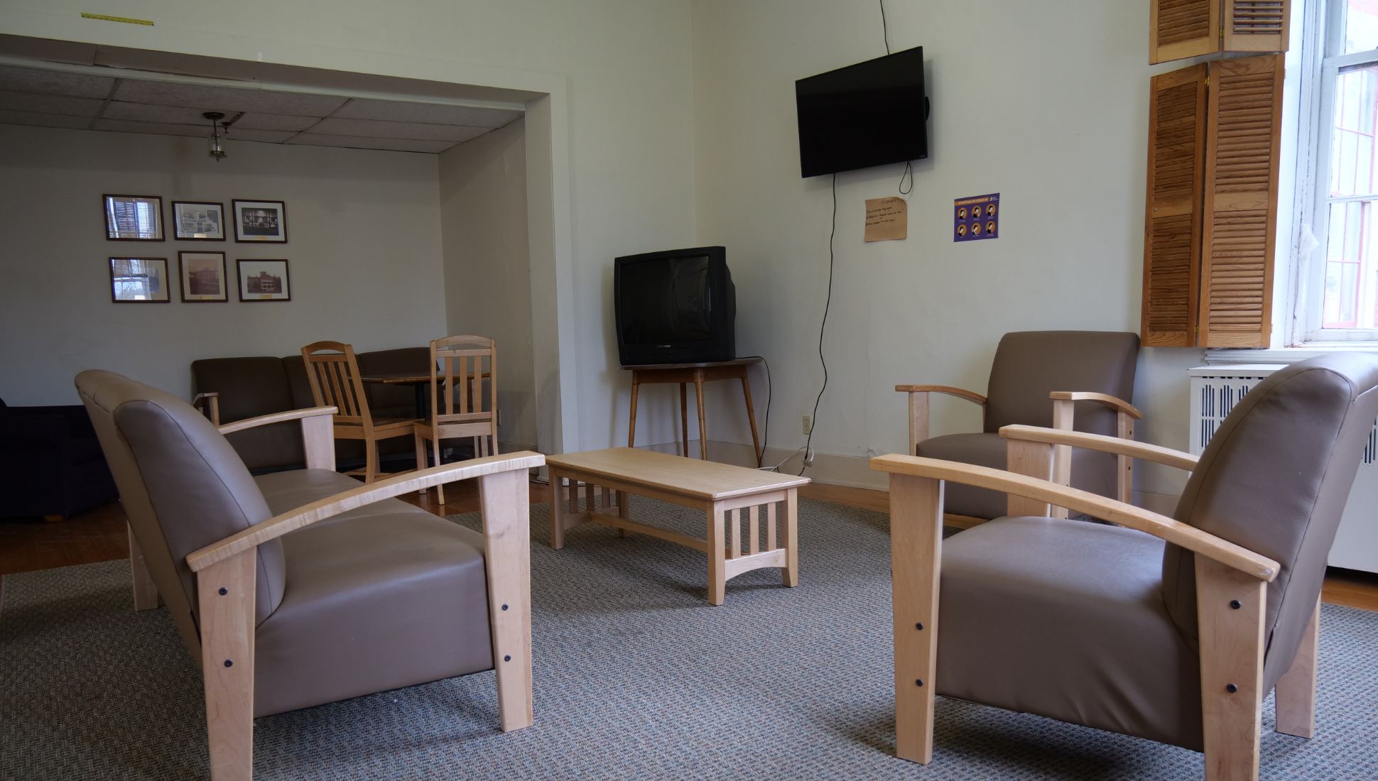 A dormitory lounge with brown cushioned chairs and a wooden coffee table arranged in a circle on a carpeted floor. A flat-screen TV is mounted on the wall with visible cords, and an older box-style TV sits on a wooden stand beneath it. A window with wooden shutters allows natural light in, and a radiator is positioned below the window. In the background, a dining nook contains a brown couch and a small wooden dining set with framed photos on the wall.