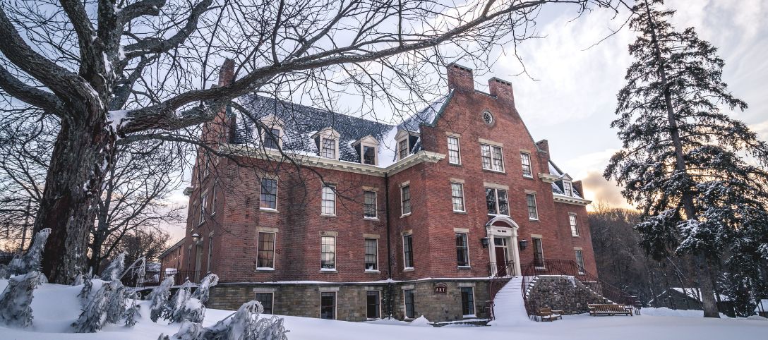Red brick residence hall at Alfred University during winter, surrounded by snow-covered trees and pathways under a soft, cloudy sky at sunset