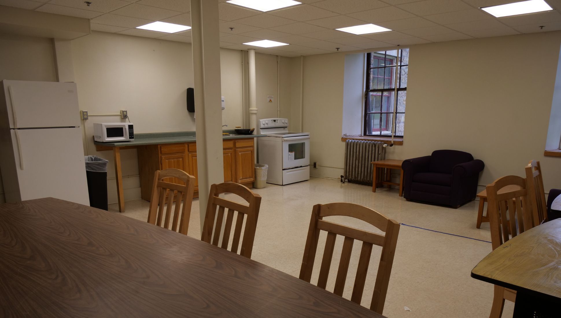 A dormitory lounge with brown cushioned chairs and a wooden coffee table arranged in a circle on a carpeted floor. A flat-screen TV is mounted on the wall with visible cords, and an older box-style TV sits on a wooden stand beneath it. A window with wooden shutters allows natural light in, and a radiator is positioned below the window. In the background, a dining nook contains a brown couch and a small wooden dining set with framed photos on the wall.