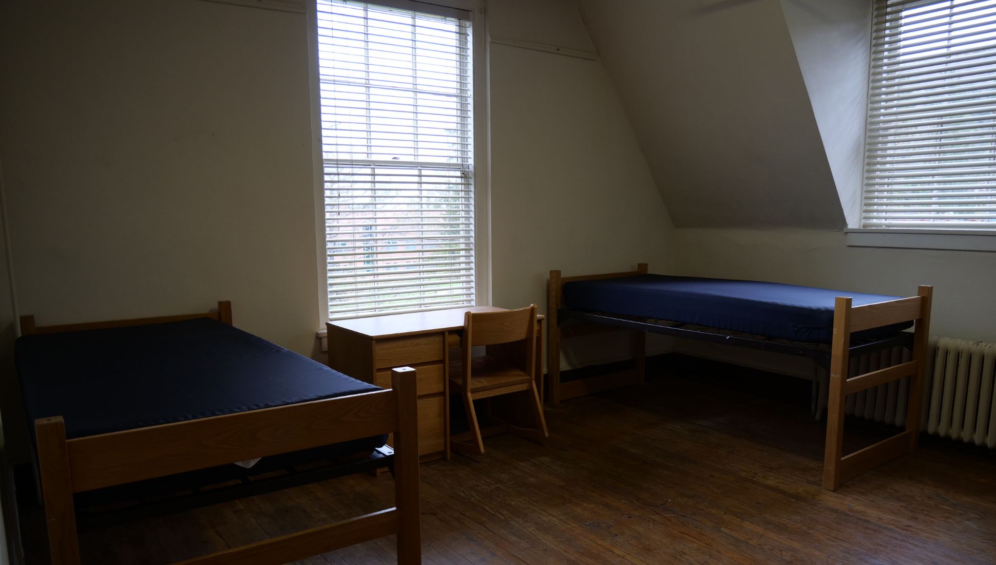 A simple college dorm room with two wooden bed frames and blue mattresses, a shared wooden desk and chair positioned between two large windows with blinds. The room has dark wooden floors, light-colored walls, and a sloped ceiling section near one window. A radiator sits beneath the right window. Natural light fills the room through the windows.