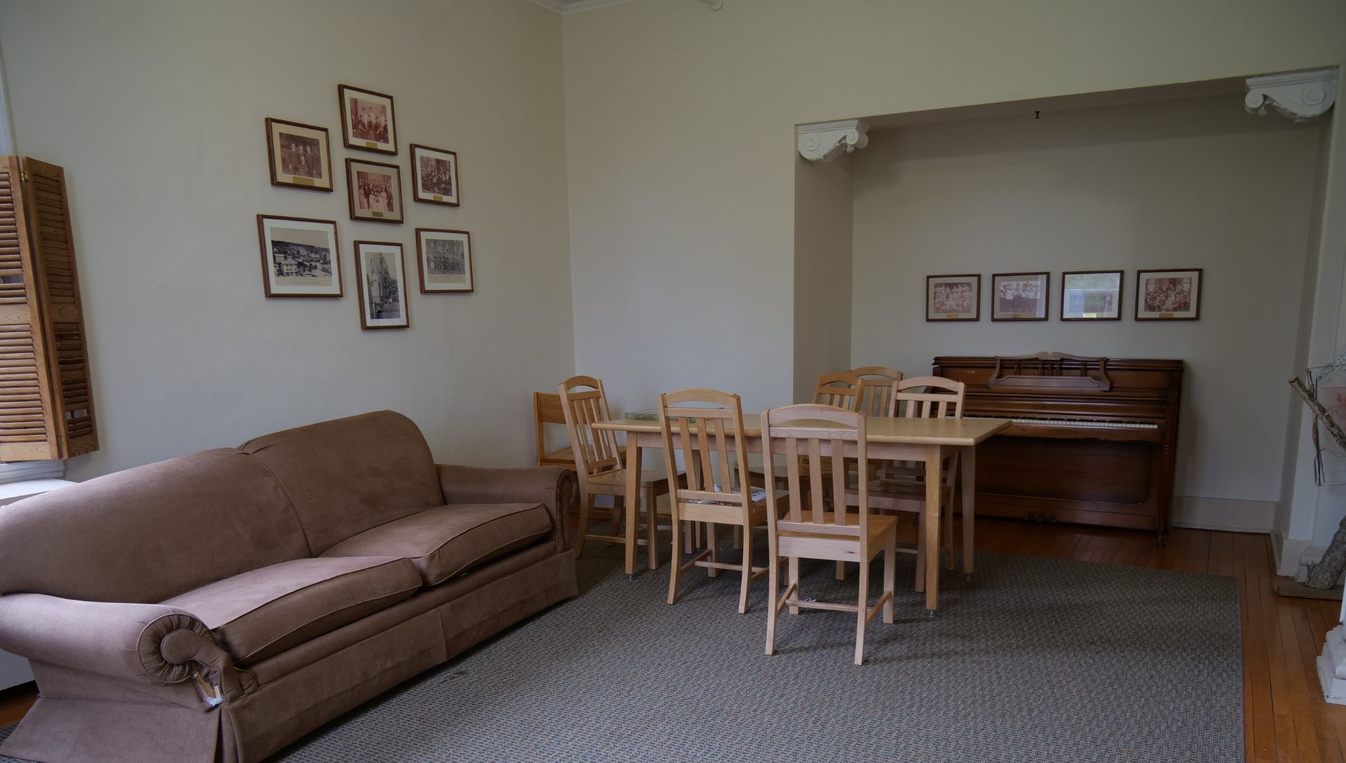A dormitory lounge area featuring a brown upholstered couch, a wooden dining table with eight chairs, and an upright piano placed in a recessed nook. Framed historical photographs decorate the walls, and a window with wooden shutters is partially visible on the left. The room has light-colored walls, hardwood flooring, and a carpeted area under the furniture. Decorative crown molding is present above the piano nook.