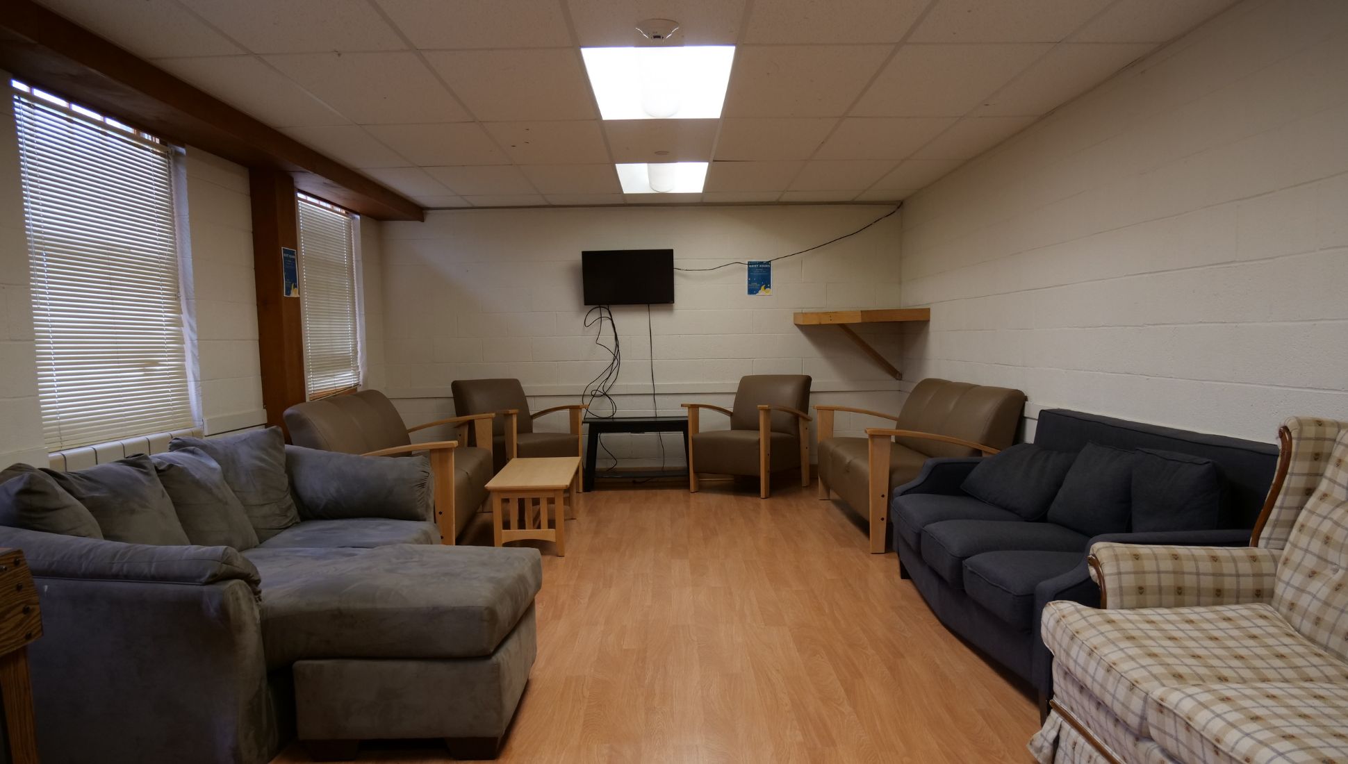 A college common room with various seating options including a gray sectional, a navy blue couch, two brown armchairs, and a plaid armchair. The room features wood-style flooring, a wall-mounted TV with visible cords, a couple of small coffee tables, and a wooden wall shelf. The walls are white-painted cinder block, and three large windows with blinds allow natural light into the space. Fluorescent ceiling lights brighten the room.