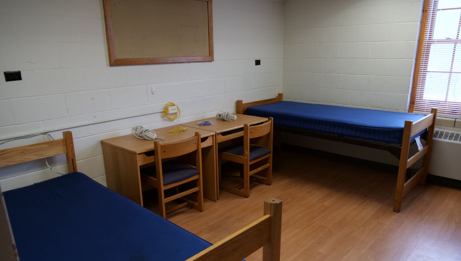 A simple college dorm room with two wooden bed frames and blue mattresses, two wooden desks with chairs, and a cork bulletin board on the wall. The desks have power strips and an ethernet cable on top. The room features light-colored cinder block walls, a large window with blinds, and wood-style flooring. A heater is located beneath the window.