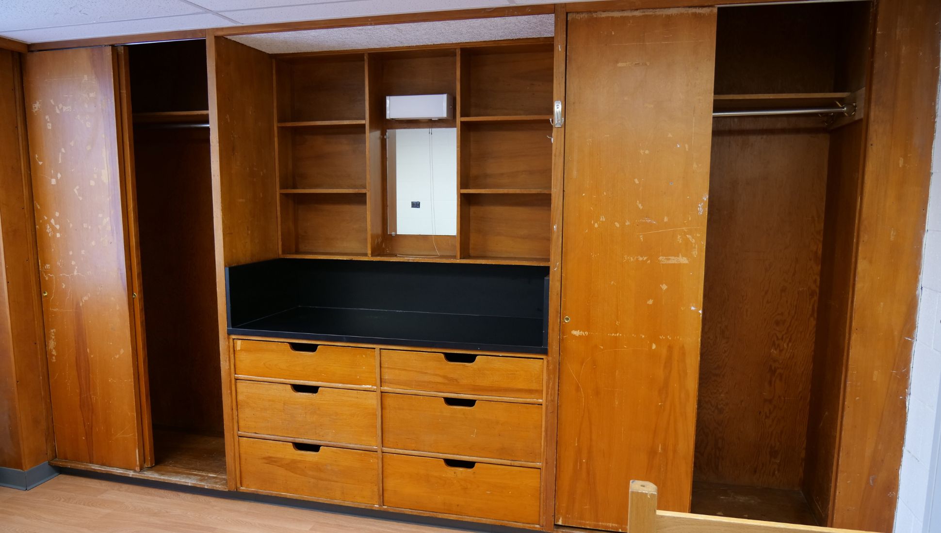 A built-in dormitory closet and storage unit made of wood, featuring multiple shelves, a mirror, six drawers, and a black countertop area. The unit has three open closet sections with clothing rods and sliding wooden doors, showing signs of wear and scratches. The floor is wood-style laminate, and part of a wooden bed frame is visible in the foreground.