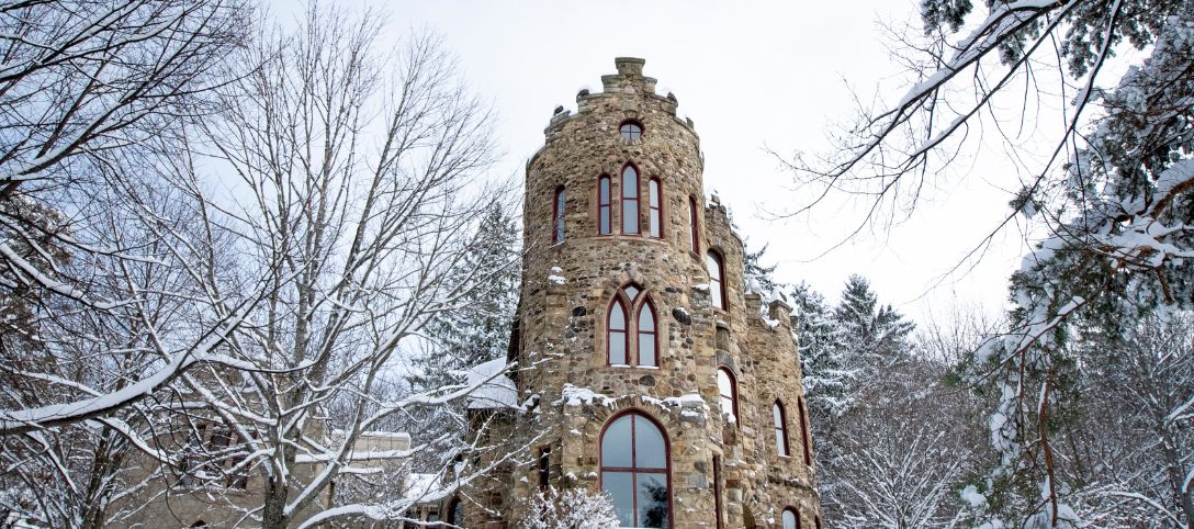 Low-angle view of the stone Allen Steinheim Castle at Alfred University, with arched windows and a rounded turret, framed by autumn trees under a bright blue sky.