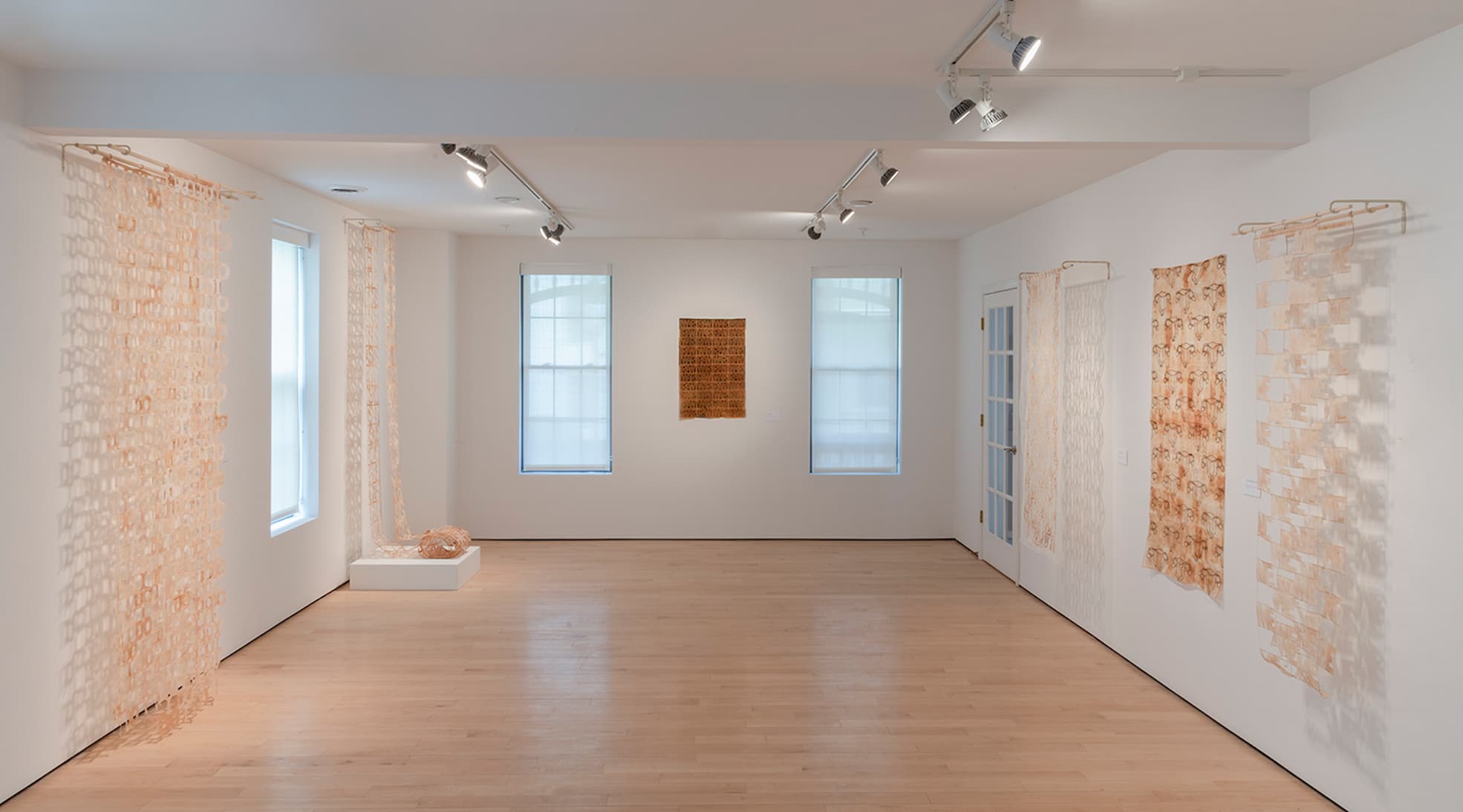 An art gallery room with light wooden floors and white walls. The room features five textile artworks hanging from the ceiling and walls. Two large, translucent fabric pieces hang on either side of a window on the left wall. In the center of the back wall, between two windows, is a smaller rectangular textile piece with intricate patterns. On the right wall, there are three more hanging textiles with varying designs and textures. The room is well-lit by track lighting on the ceiling.