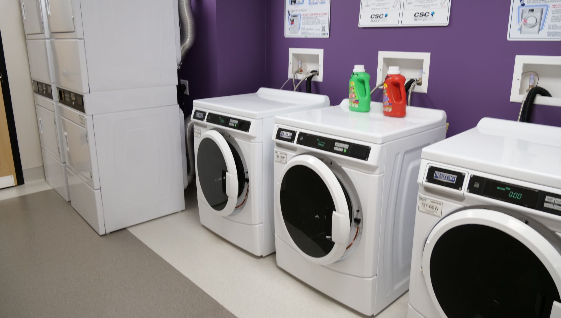 A dormitory laundry room with three front-loading washing machines and a set of stacked dryers against a purple wall. Two bottles of laundry detergent, one green and one red, sit on top of a washer. Instructions for using the machines are posted on the wall above. The floor is a mix of gray and white tile.
