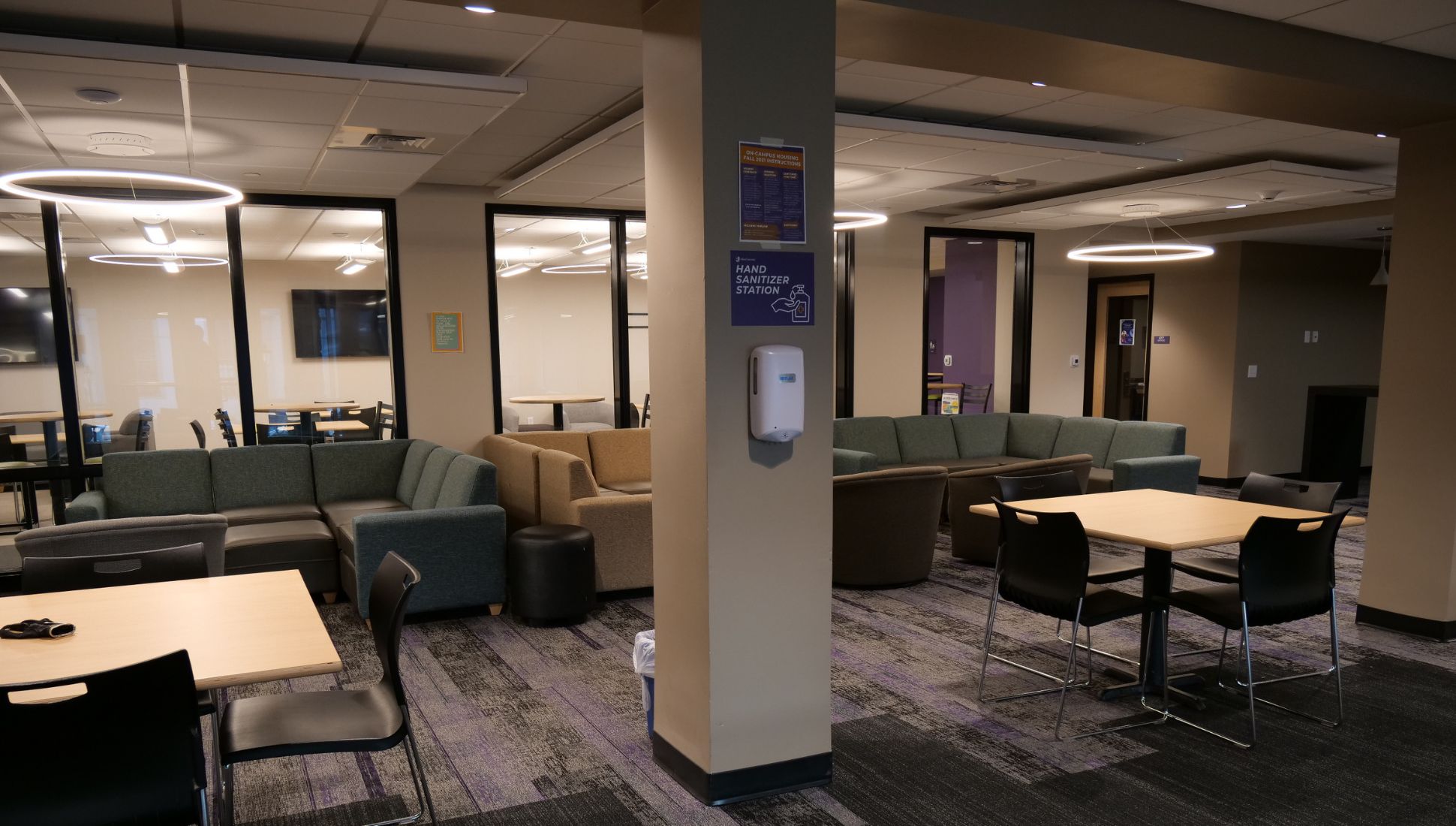 A cozy student lounge with sectional sofas in green and neutral tones, small round ottomans, and square wooden tables with black chairs. A hand sanitizer dispenser is mounted on a central column labeled "Hand Sanitizer Station." The room features glass-walled study rooms in the background, modern circular ceiling lights, and a carpeted floor with purple and gray patterns.