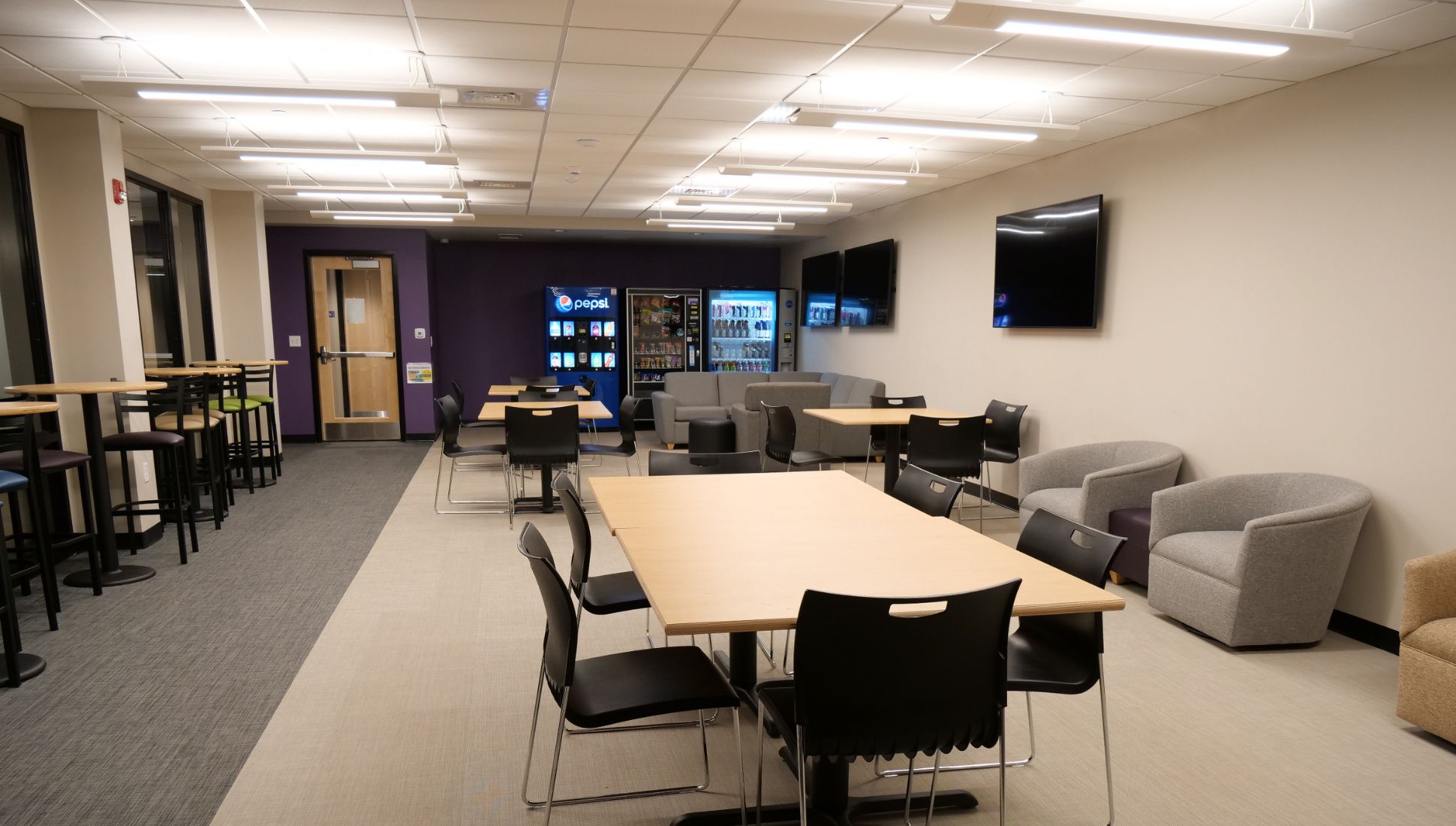 A modern college lounge with multiple tables and chairs, gray lounge chairs, and vending machines including a Pepsi machine in the back. There are three large flat-screen TVs mounted on the wall, and the space is well-lit with ceiling lights. High-top tables with colorful stools are placed along the left wall, and the room has a mix of carpet and tile flooring.