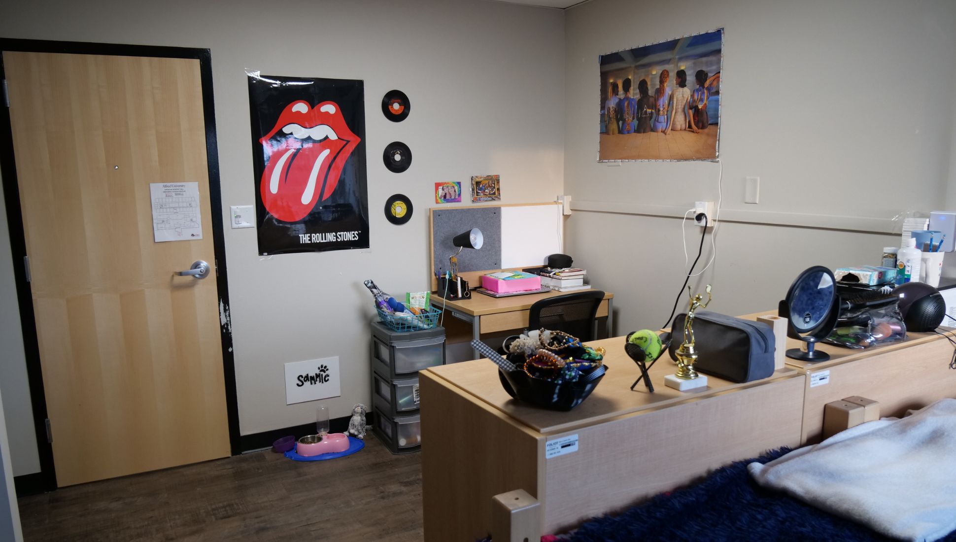 A college dorm room with a light wood door, a Rolling Stones poster, and several vinyl records decorating the wall. There's a small study desk with a lamp, books, and storage bins beside it. A personalized pet area on the floor includes a food and water bowl labeled "Sammie." On the dresser are a trophy, mirror, and personal care items. The room has a cozy setup with wood-style flooring and posters adding a personal touch.
