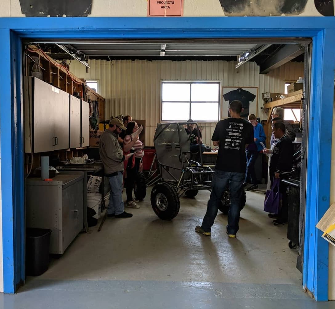 Inside a workshop marked 'PROJECTS AREA,' several people are gathered around a large wheeled vehicle in progress. The garage-like space features tools, cabinets, and workbenches along the walls. Some individuals appear to be discussing or examining parts of the vehicle, while others look on. The atmosphere suggests a collaborative, hands-on learning or engineering environment.