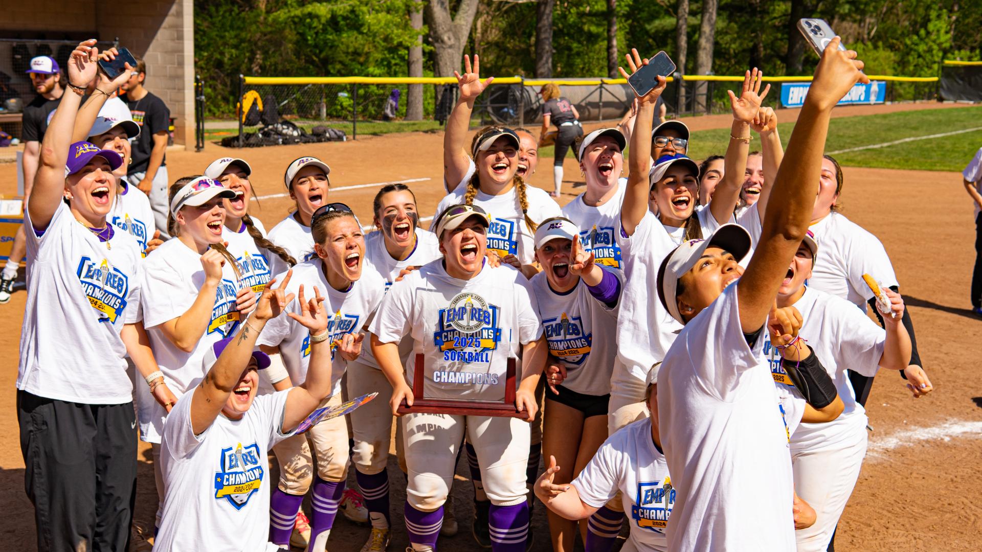 A women's softball team wearing white shirts poses together on a field, holding a championship trophy and taking a group selfie.