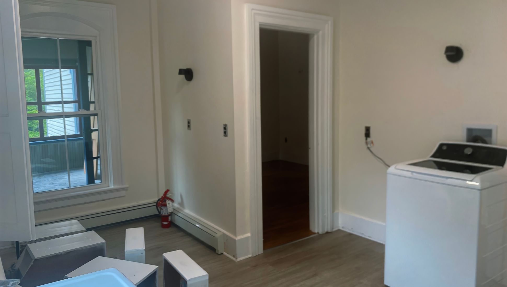 A narrow stairwell with white walls leading down to an open blue door marked by a red "EXIT" sign above. The stairs show signs of wear, and a small, partially detached electrical box or outlet dangles near the bottom right. Outside the door, a wooden porch with lattice fencing is visible, along with green grass beyond.