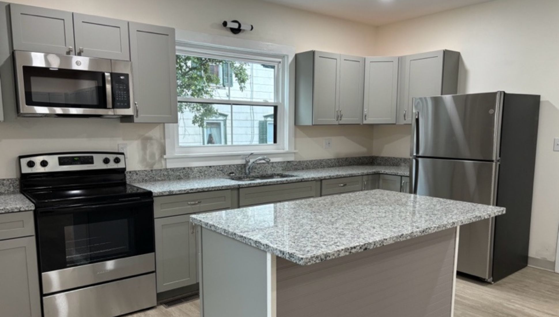 Modern kitchen with gray cabinets, granite countertops, a central island, and stainless steel appliances including a microwave, oven, and refrigerator. A window above the sink provides natural light.