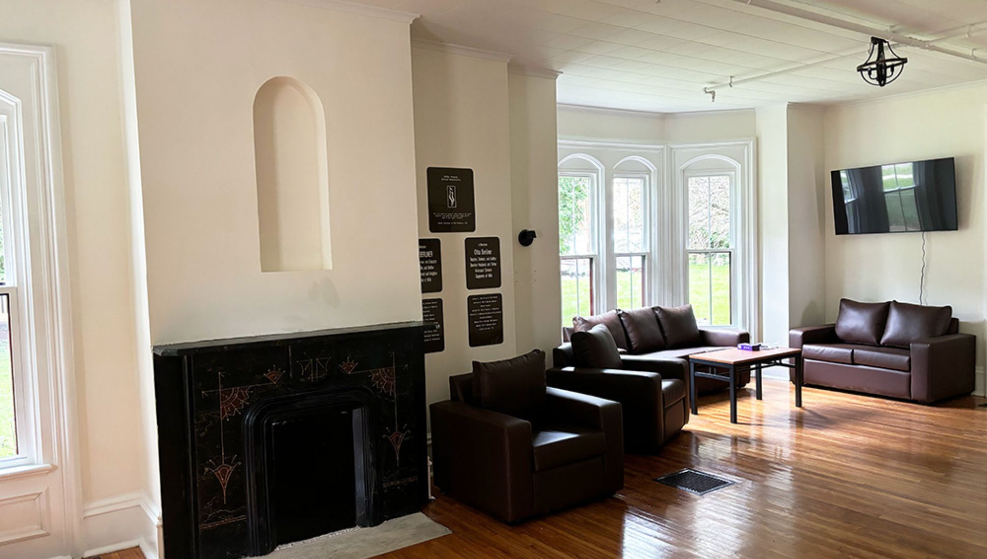 Bright, spacious living room with large windows, brown leather sofas and armchairs, a wall-mounted TV, and a decorative fireplace. Hardwood floors and light-colored walls create a warm, welcoming atmosphere.