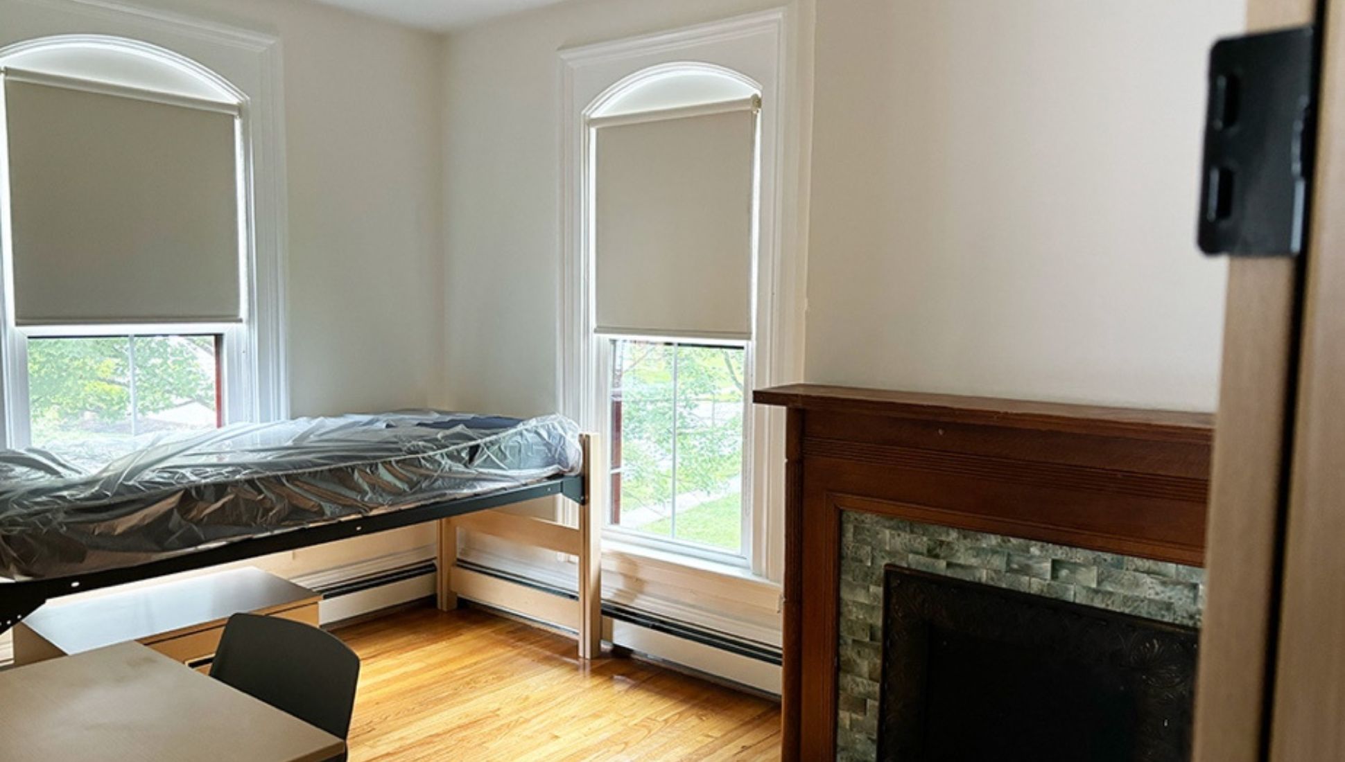 Simple, well-lit bedroom with hardwood floors, two tall windows with shades, a single bed frame with mattress, a wooden desk and chair, and a decorative fireplace with a wooden mantel.