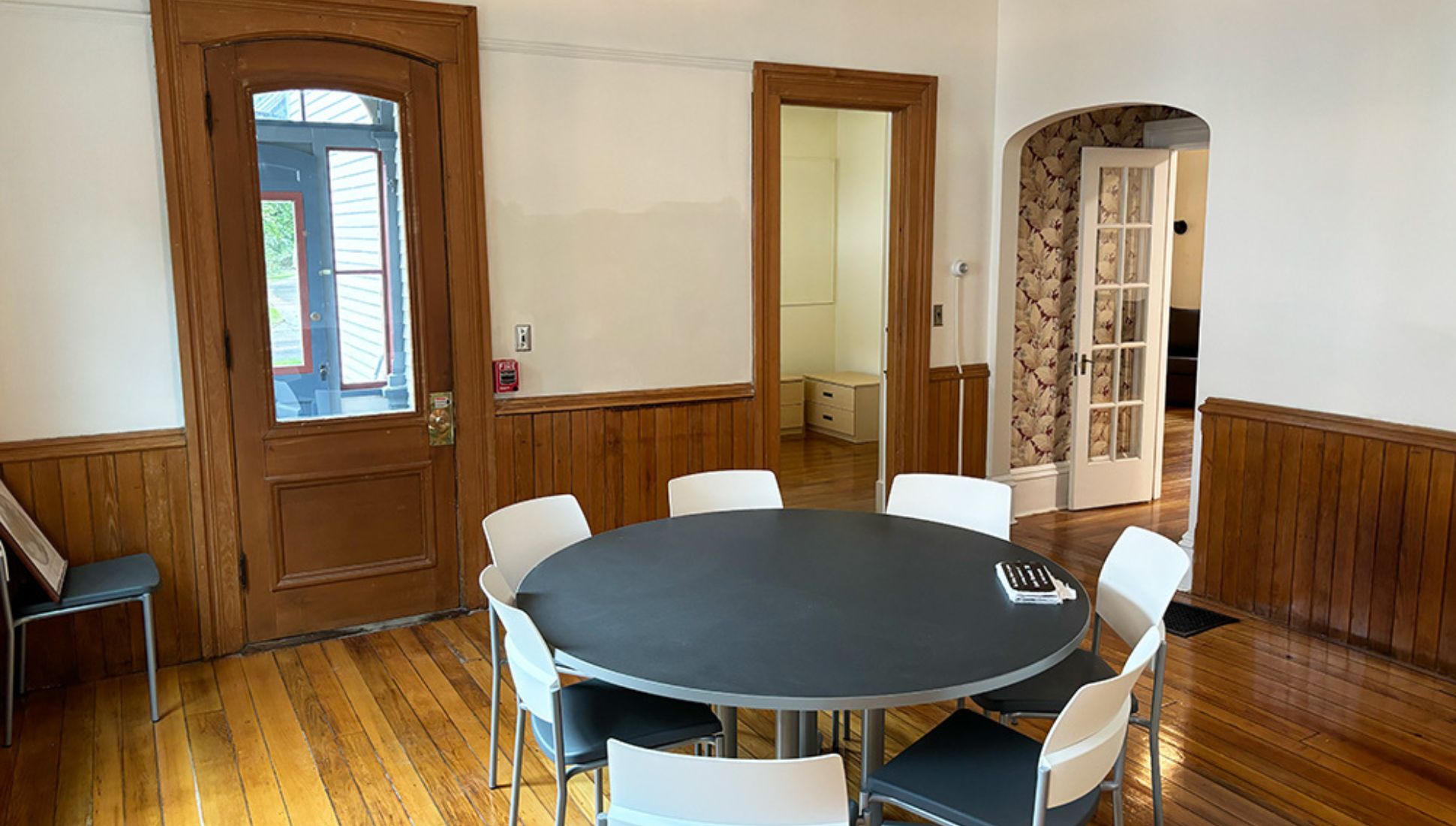Cozy dining or meeting area with a round black table surrounded by white chairs, hardwood floors, wood-paneled lower walls, and several doorways leading to adjacent rooms, including one with floral wallpaper and a glass-paned door.