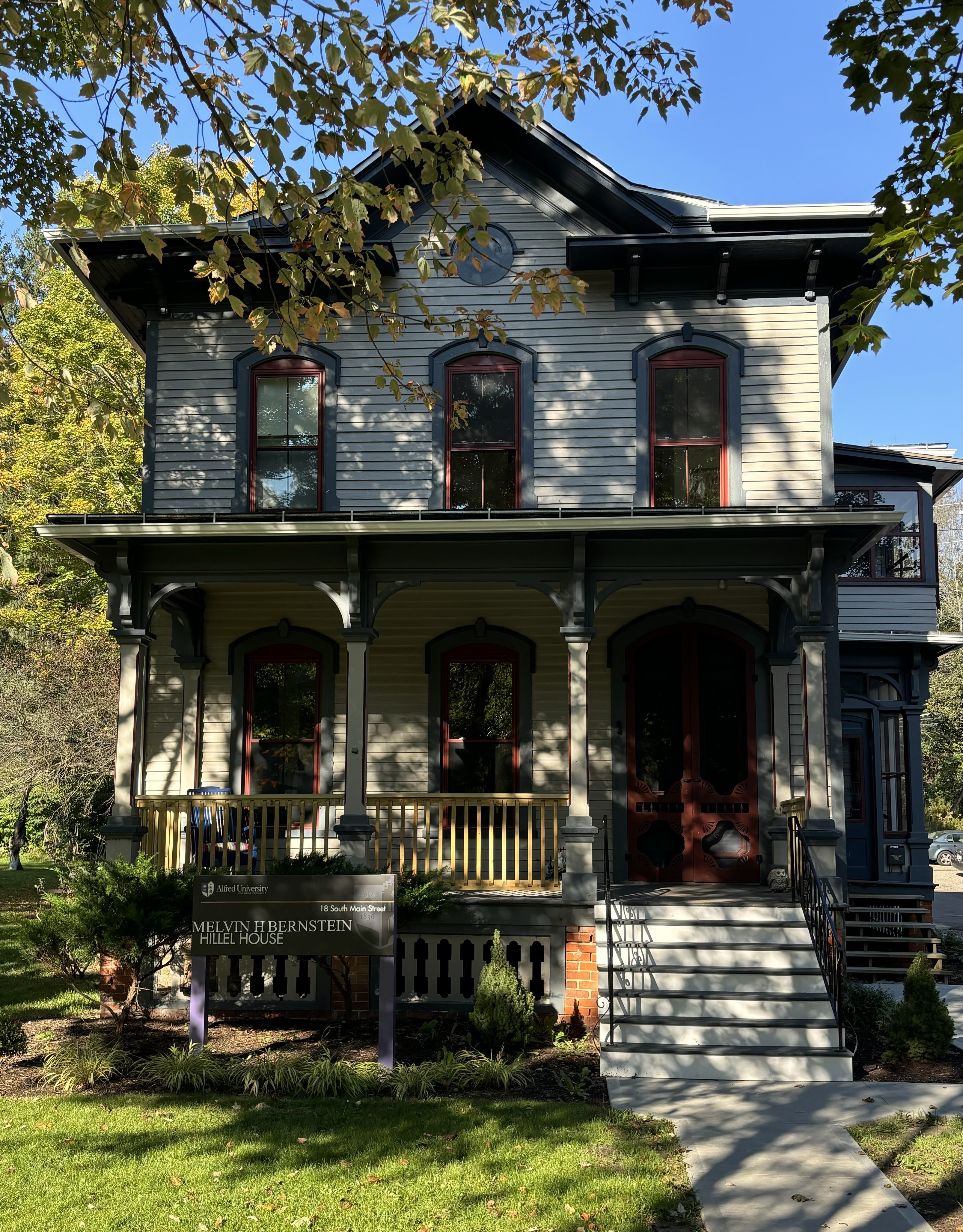 Front view of the Melvin H. Bernstein Hillel House, a two-story historic-style building with gray siding, dark trim, red-accented windows, and a covered porch. Surrounded by trees and a grassy lawn with a concrete walkway leading to the steps.