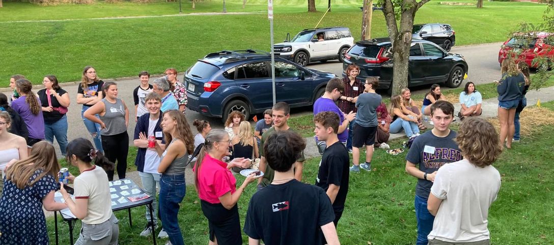 Students gather on the lawn outside the Alfred University Honors House during a social event, chatting, smiling, and enjoying food in a relaxed outdoor setting surrounded by trees and parked cars.