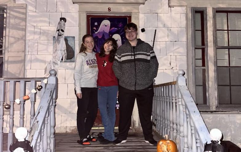 Three Alfred University students smile and pose on the decorated front porch of the Honors House during Halloween, surrounded by spooky decorations including a ghost door cover, hanging skeleton, string lights, and a carved pumpkin.