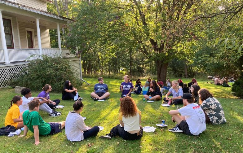 Alfred University students sit in a large circle on the grass outside the Honors House, enjoying a sunny day while sharing food and conversation in a relaxed outdoor setting surrounded by trees.