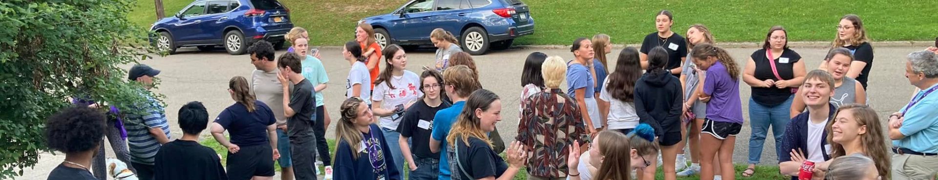 A large group of Alfred University students and faculty gather outside on a sidewalk and grassy area during an Honors House welcome event, chatting and mingling near a table with name tags.