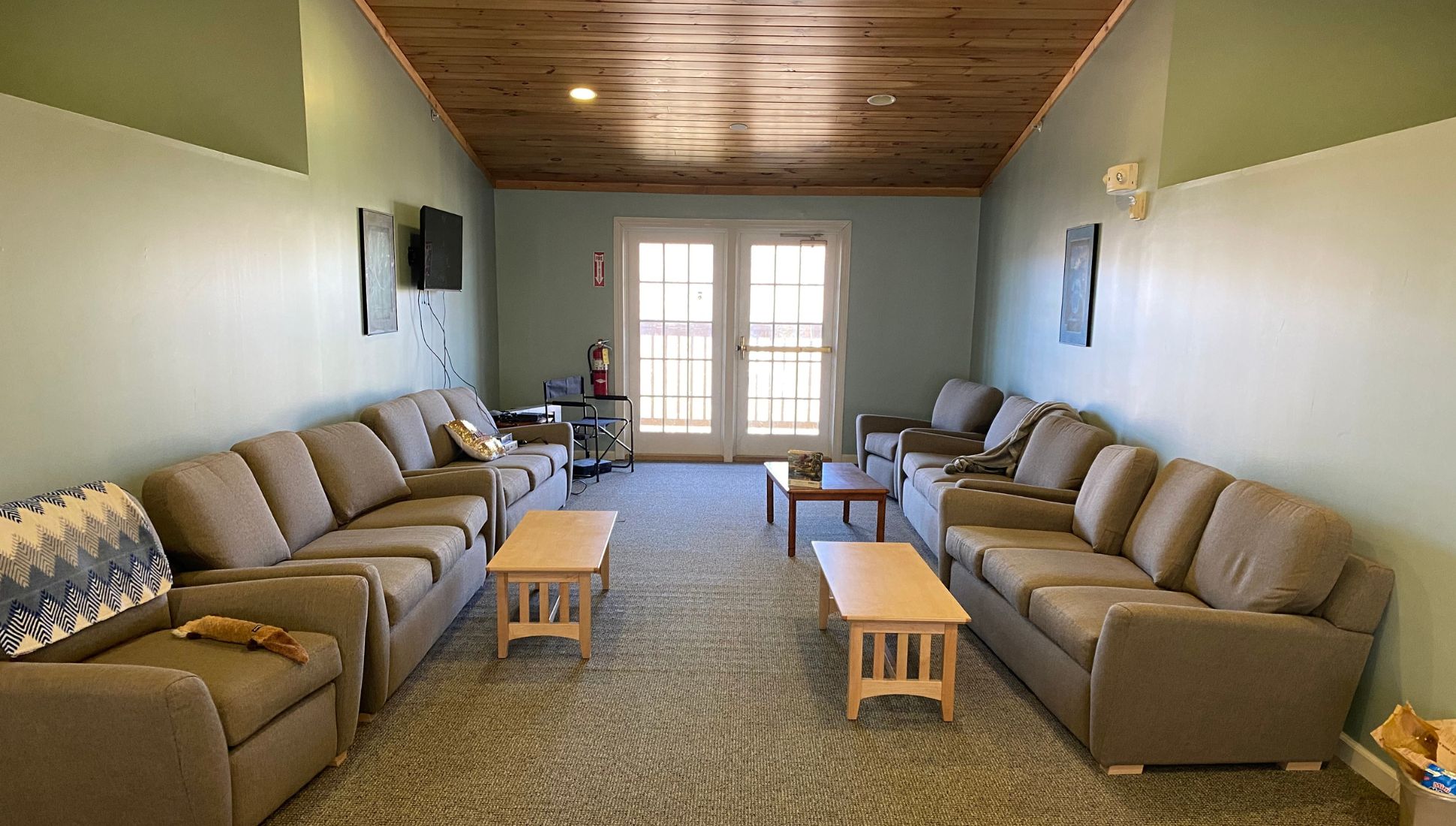 A cozy dormitory lounge with soft green walls, a wooden plank ceiling, and carpeted flooring. The room is furnished with multiple gray couches and armchairs arranged in two rows facing each other, with three wooden coffee tables in between. A wall-mounted TV and framed artwork decorate the space, and a fire extinguisher sits near a pair of French doors at the far end of the room. Blankets and throw pillows add a homely touch to the seating.