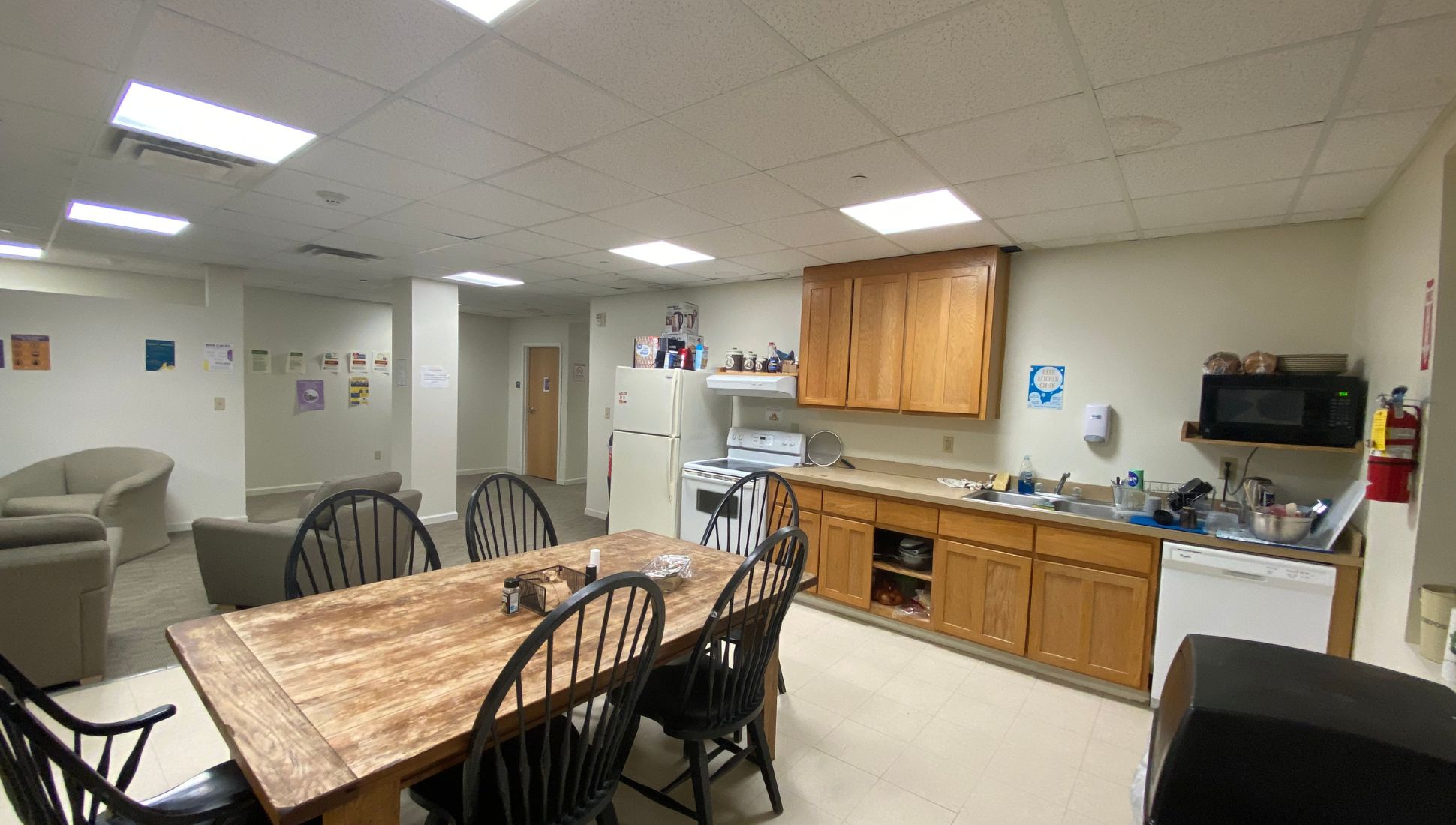 A communal kitchen and lounge area with wooden cabinetry, a refrigerator, oven, microwave, dishwasher, and sink. A rustic wooden dining table with black chairs sits in the center. Various kitchen items are on the counters and shelves, including dishware, cooking utensils, and snacks. In the background, beige armchairs form a seating area against a wall decorated with colorful posters and announcements. The room has tiled floors, drop ceiling lights, and a cozy, lived-in feel.
