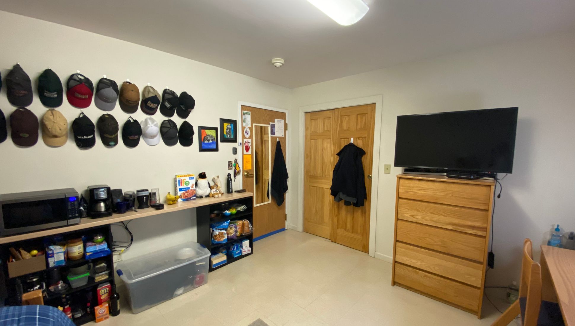 A neatly organized dorm room with a wooden dresser holding a large flat-screen TV, a mini kitchen setup with a microwave, coffee maker, and snacks on a shelf, and a collection of baseball caps displayed on the wall. The room has white walls, a tiled floor, and a wooden door with a mirror and coat hanging on it. There's also a plastic storage bin under the shelf and a wooden desk partially visible in the lower right corner. A board game and plush toys sit on the counter, adding a personal touch.