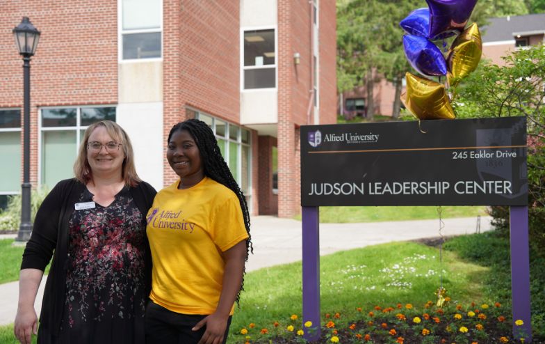 Two smiling people standing in front of the Judson Leadership Center sign at Alfred University, with colorful balloons and a flower bed decorating the entrance.